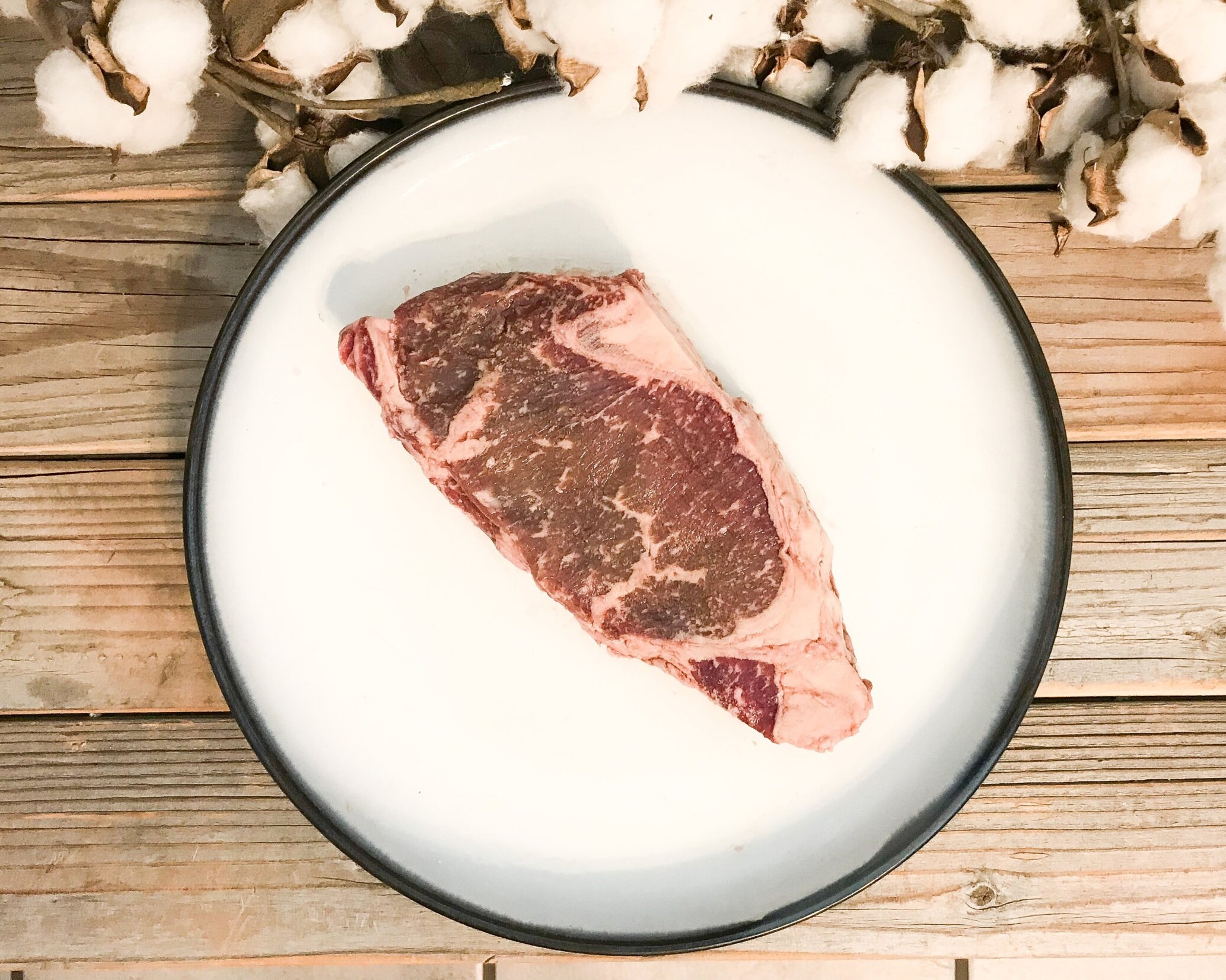 Raw ribeye steak on a white plate, surrounded by cotton bolls, set against a rustic wooden background, showcasing marbling and texture for culinary presentation.