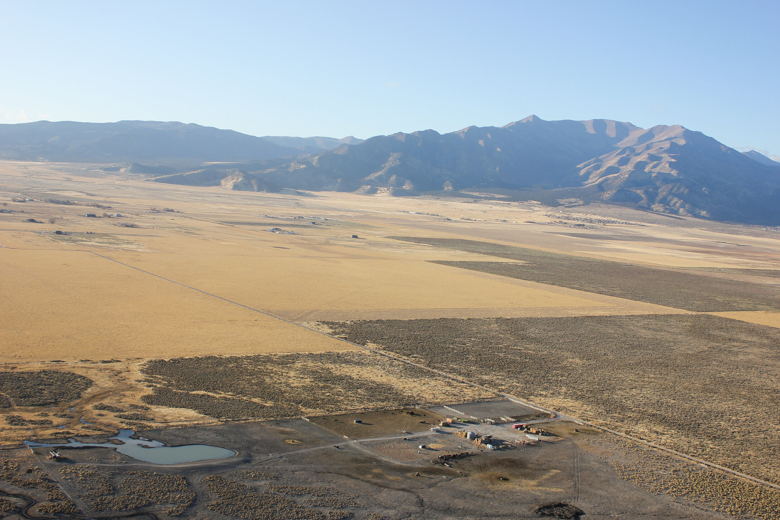 Christiansens+Family+farm+Utah+-+IMG_4698 Aerial view of a vast, arid landscape featuring a small settlement near a pond, surrounded by golden grasslands and mountains in the background. The image captures the expansive terrain of the area, highlighting natural features and human habitation.