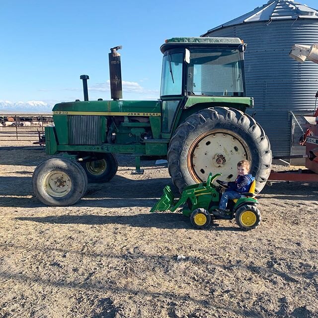 image-asset - 2026-07-17T142710.643 A young child plays with a toy tractor next to a large green John Deere tractor on a farm, with a silo and clear blue sky in the background.