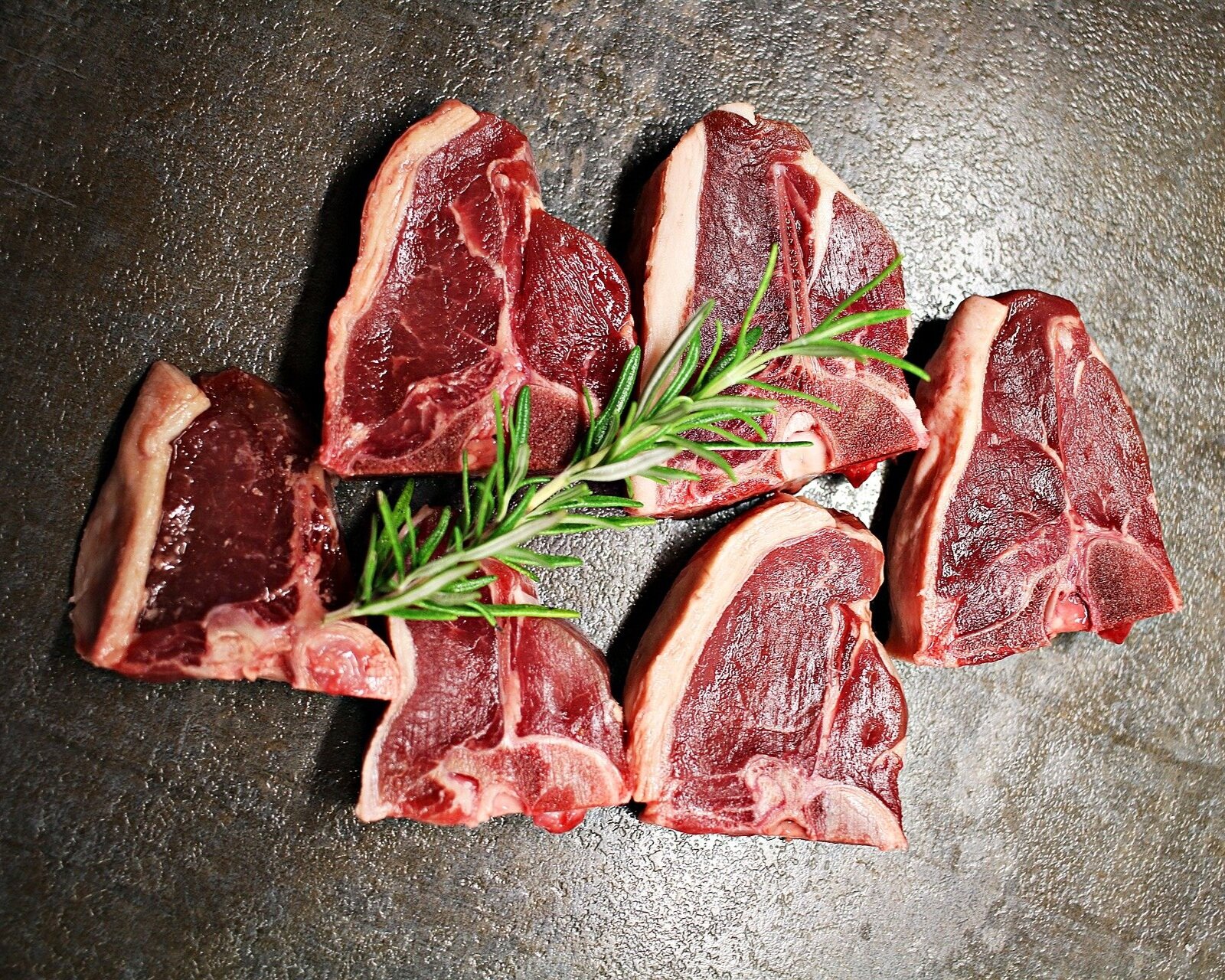 Fresh cuts of meat arranged on a dark surface, garnished with a sprig of rosemary, showcasing high-quality beef steaks ideal for grilling or roasting.